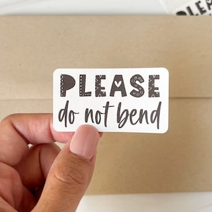 May include: A white sticker with black text that reads "PLEASE do not bend". The sticker is being held by a hand.