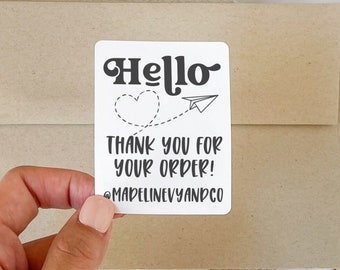 May include: A white sticker with the word "Hello" in a black, retro font, a heart, and a paper airplane. Below, it reads "Thank you for your order!" and the seller's Instagram handle. The sticker is held over a brown envelope and a notebook.