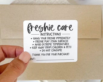May include: A hand holding a white sticker with black text that reads "freshie care" and instructions. The sticker is on a brown envelope. Other items include gold and pink paper clips, a tassel, and a ribbon.