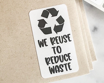 May include: White rectangular stickers with a black recycling symbol and the text "WE REUSE TO REDUCE WASTE". The stickers are on a roll and are placed on top of a stack of brown paper.