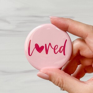 May include: Pink round button with the word "loved" in pink script with a small pink heart in front of the "v".