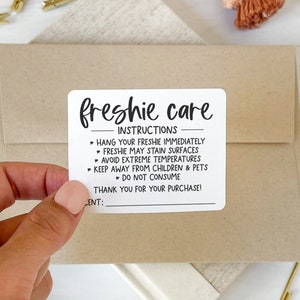 May include: A hand holding a white sticker with black text that reads "freshie care" and instructions. The sticker is on a brown envelope. Other items include gold and pink paper clips, a tassel, and a ribbon.