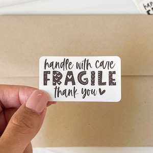 May include: White sticker with black text that reads "handle with care FRAGILE thank you" with a small heart symbol. The sticker is on a brown paper background.