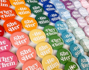 Può includere: Una collezione di bottoni rotondi colorati con diverse combinazioni di pronomi, tra cui "He Him", "She Her" e "They Them".