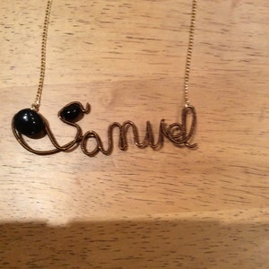 Op de afbeelding: Een gouden ketting met de naam "Samuel" gespeld in draad met zwarte kralen aan beide uiteinden.