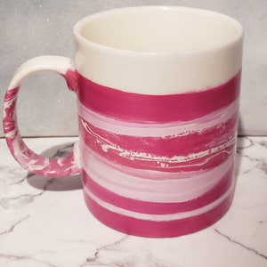 May include: A white ceramic mug with pink and white horizontal stripes. The mug has a handle and is sitting on a white marble surface.