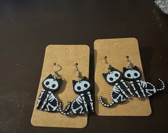 May include: Two pairs of black and white skeleton cat earrings on brown card stock. The earrings are dangle style with silver hooks.