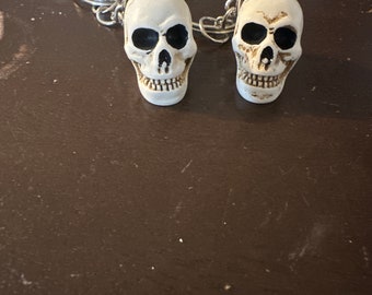 May include: Two white skull keychains with black eye sockets and a silver chain. The keychains are on a brown surface.