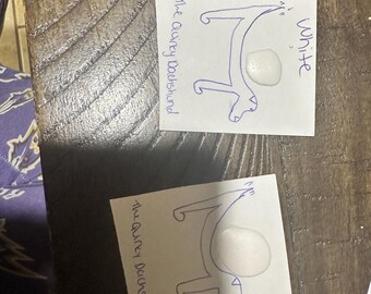 May include: Two white cards with a blue outline drawing of a dachshund and the text "The Quirky Dachshund". One card has the word "White" written in blue. A white, oval-shaped object is on each card.