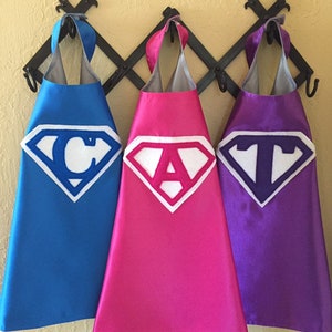 May include: Three superhero capes hanging on a rack. The capes are blue, pink, and purple. Each cape has a white felt Superman symbol with a letter in the center. The letters spell out "CAT".