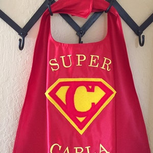 May include: Red superhero cape with a yellow Superman logo and the name "SUPER CARLA" embroidered in yellow.