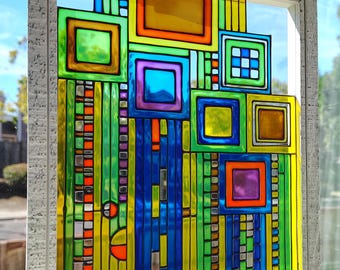 May include: Abstract stained glass window art in a grey frame. The design features colourful squares and rectangles in shades of blue, green, yellow, orange, and purple. The artwork is displayed in front of a window, allowing natural light to pass through.