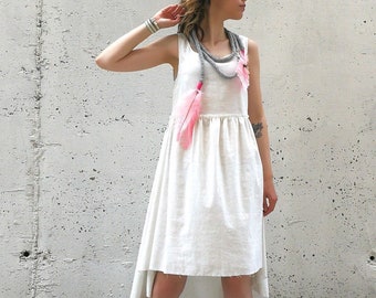 May include: A white linen dress with a high low hemline and a gray and pink feather necklace. The dress has a gathered waist and a sleeveless design. The model is wearing white sneakers.