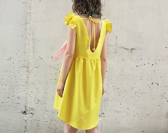 May include: A yellow sleeveless dress with a deep V-back and ruffled straps. The dress has a loose fit and a slightly shorter hem in the back. The model is wearing white sneakers.