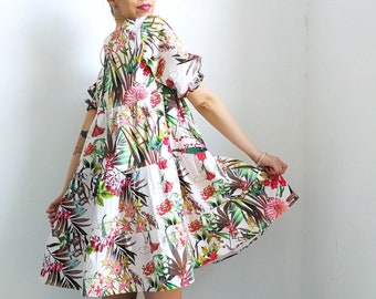 May include: A white dress with a floral print featuring tropical leaves and flowers. The dress has a loose fit and a short, ruffled hem. The model is wearing white sneakers.