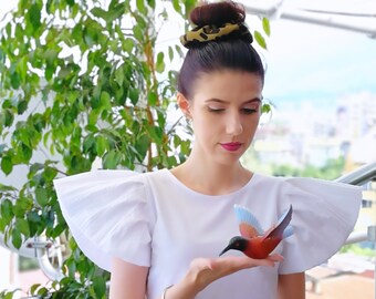 May include: A woman wearing a white blouse with large, ruffled sleeves and a leopard print skirt is holding a small, red, blue, and white hummingbird figurine.