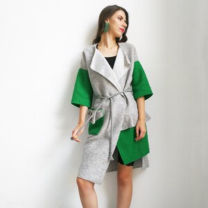 May include: A gray and green long-sleeved cardigan with a tie belt. The cardigan has a long, asymmetrical hemline and a large pocket on the front. The model is wearing black boots.