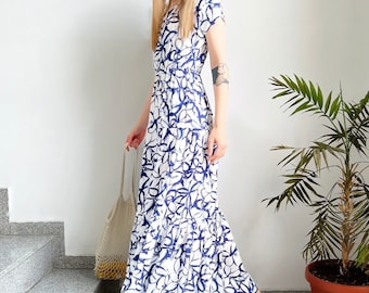 May include: A white maxi dress with a blue floral print. The dress has a fitted bodice and a flowing skirt. The model is wearing the dress with black sandals.