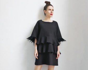 May include: A black dress with a ruffled skirt and feather trim on the sleeves. The dress is knee-length and has a loose fit. The model is wearing black platform sandals.