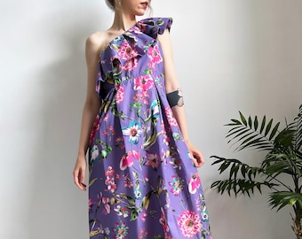 May include: A long, purple maxi dress with a floral print. The dress has a one-shoulder design with a ruffle detail.