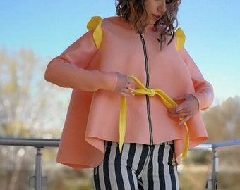 May include: A woman wearing a peach-colored jacket with yellow accents and black and white striped pants. She is standing on a balcony and wearing yellow high-top sneakers.