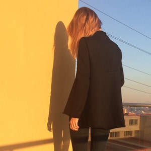 May include: A woman wearing a black blazer, blue jeans, and black ankle boots stands in front of a yellow wall. Her shadow is cast on the wall behind her.