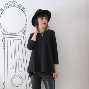 May include: A woman wearing a black hat, black top, and black leather pants. She is standing in front of a white wall with a black and white clock on it.