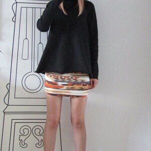May include: A woman wearing a black turtleneck sweater, a black fedora hat, and a colorful patterned mini skirt. She is standing in front of a white wall with a black and white clock illustration.