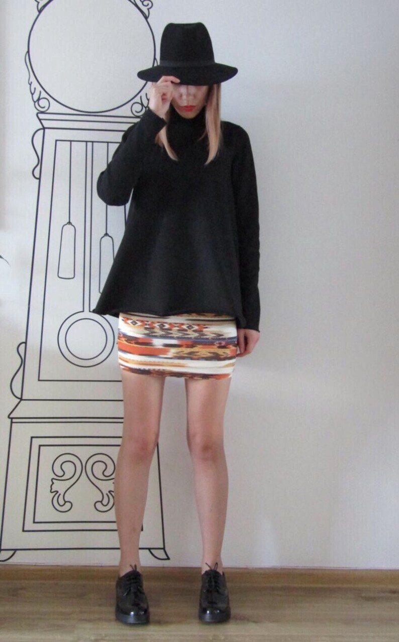 May include: A woman wearing a black turtleneck sweater, a black fedora hat, and a colorful patterned mini skirt. She is standing in front of a white wall with a black and white clock illustration.