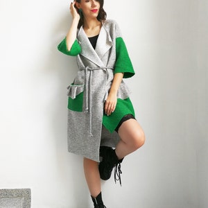 May include: A gray and green cardigan with a long, flowing design. The cardigan has a tie belt and a large pocket on the left side. The model is wearing black boots.
