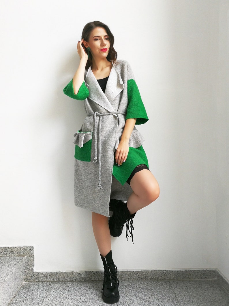 May include: A gray and green cardigan with a long, flowing design. The cardigan has a tie belt and a large pocket on the left side. The model is wearing black boots.