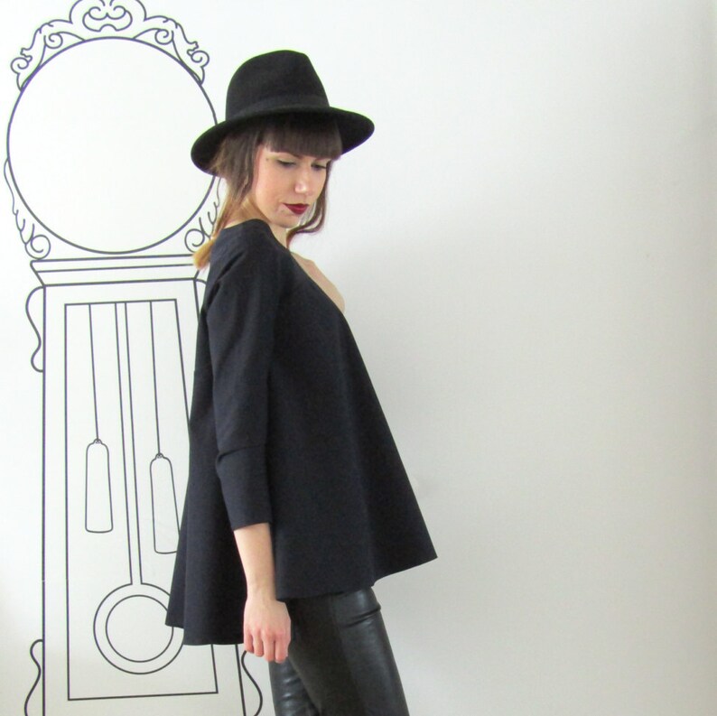 May include: A woman wearing a black, wide-collared,  long-sleeved top with a slightly flared hem. She is wearing black pants and a black fedora.