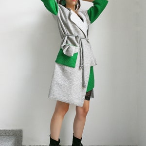 May include: A gray and green cardigan with a white collar and a tie belt. The cardigan has a large patch pocket on the front. The model is wearing black boots.