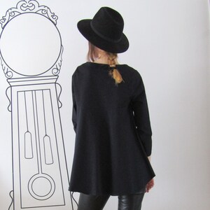 May include: A black long-sleeved tunic with a wide, flowing hemline. The tunic is worn with black pants and a black fedora hat.