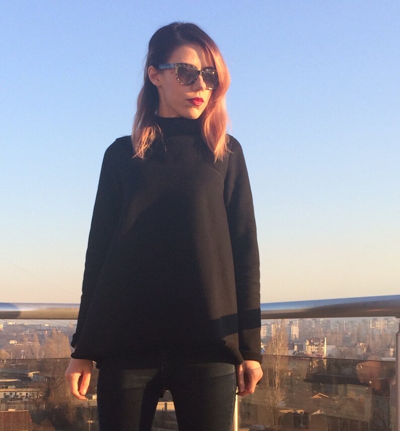 May include: A woman wearing a black mock neck shirt and blue jeans. She is wearing sunglasses and has pink hair.