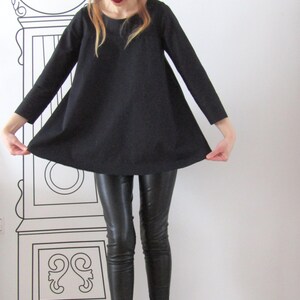 May include: A woman wearing a black, loose-fitting tunic top with long sleeves, black leather pants, and black ankle boots. She is also wearing a black fedora hat.