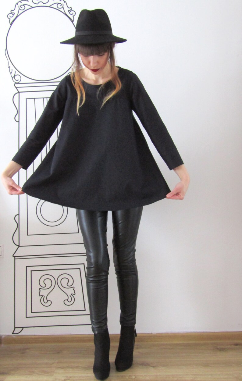 May include: A woman wearing a black, loose-fitting tunic top with long sleeves, black leather pants, and black ankle boots. She is also wearing a black fedora hat.