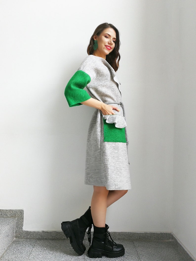 May include: A gray and green knitted dress with a ruffled pocket and a belt. The dress has long sleeves with green cuffs. The model is wearing black combat boots.