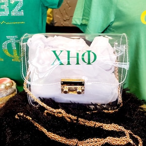 Può includere: Una borsa a tracolla trasparente in plastica con una catena dorata e una chiusura a lucchetto dorata. La borsa ha una fodera bianca e le lettere "XHQ" sono stampate in verde sul davanti.
