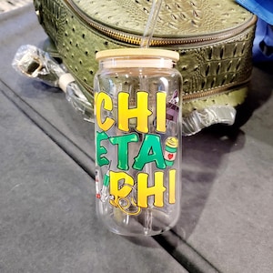 May include: A clear glass water bottle with a bamboo lid and a green and yellow design that says "CHI ETA PHI". The bottle has a straw in it.