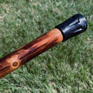 May include: A wooden walking stick with a black rubber tip. The stick is made of a light brown wood with dark brown and reddish-brown grain patterns. The tip is a solid black color.