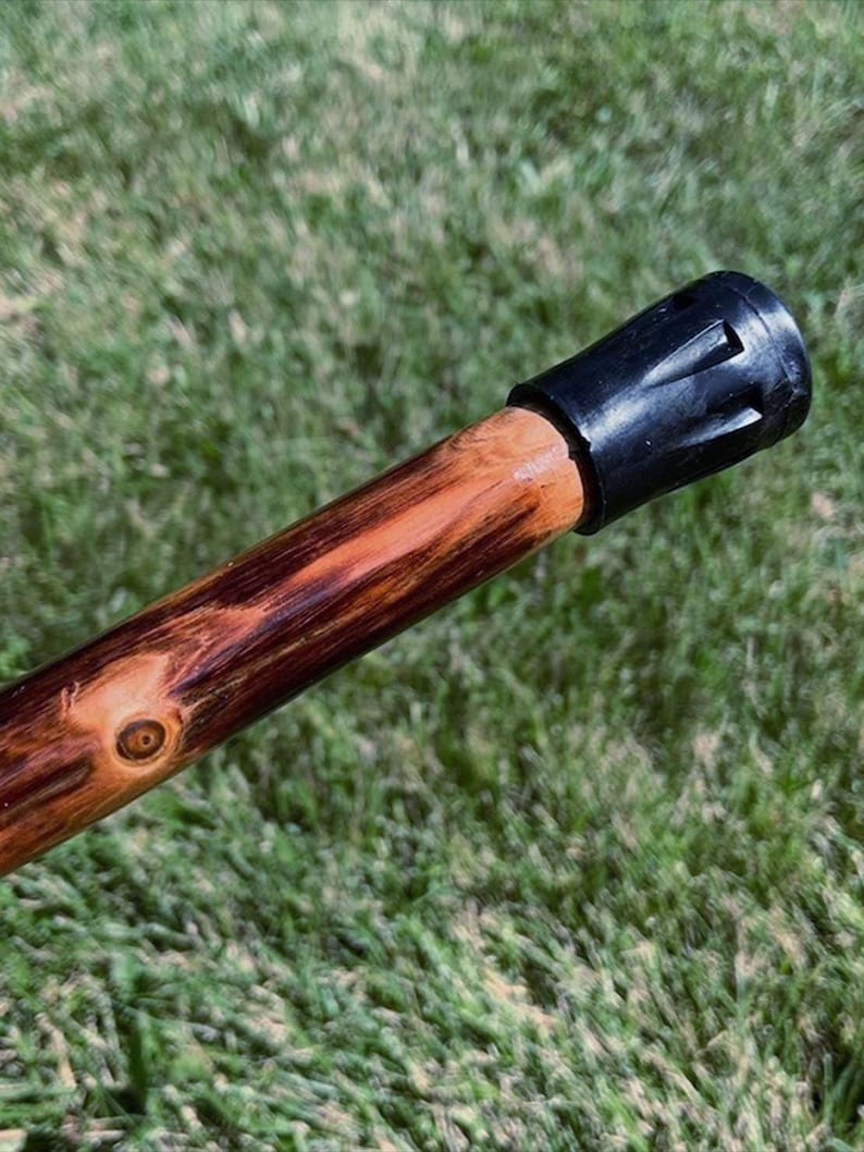 May include: A wooden walking stick with a black rubber tip. The stick is made of a light brown wood with dark brown and reddish-brown grain patterns. The tip is a solid black color.
