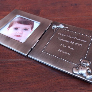 Könnte beinhalten: Ein offener, bronzefarbener Metall-Fotorahmen, der ein Babyfoto und eingravierte Details zeigt. Die rechte Seite zeigt den Namen "Abagail Marie Chambers", das Geburtsdatum, das Gewicht (3,4 kg) und die Größe (55,88 cm), mit einem Teddybären.