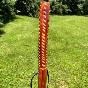 May include: A handcrafted wooden walking stick with a rich, reddish-brown finish. The upper portion features a carved pattern of elongated ovals. A black leather loop is attached near the top. The background is a green grassy area.