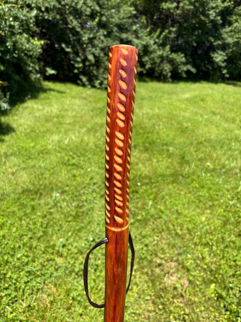 May include: A handcrafted wooden walking stick with a rich, reddish-brown finish. The upper portion features a carved pattern of elongated ovals. A black leather loop is attached near the top. The background is a green grassy area.