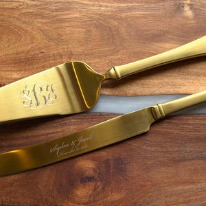 Könnte beinhalten: Goldfarbenes Tortenheber- und Messerset. Der Tortenheber hat ein eingraviertes Monogramm, und das Messer ist mit Namen und einem Datum beschriftet. Das Set liegt auf einer Holzoberfläche mit weißem Akzent.