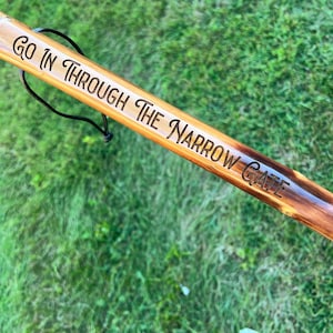 May include: A wooden walking stick with the engraved text "Go In Through The Narrow Gate." The stick is light brown with darker brown accents and a black cord attached. The background is green grass.