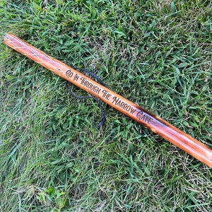 May include: A wooden walking stick with a brown finish, featuring the engraved text "GO IN THROUGH THE NARROW GATE." The stick has a decorative diamond pattern near the top and a black cord for carrying. It rests on a bed of green grass.