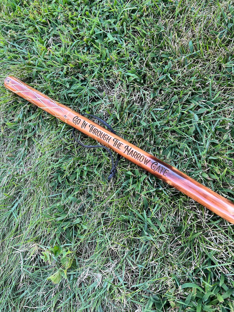 May include: A wooden walking stick with a brown finish, featuring the engraved text "GO IN THROUGH THE NARROW GATE." The stick has a decorative diamond pattern near the top and a black cord for carrying. It rests on a bed of green grass.