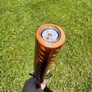 May include: A wooden walking stick with a compass embedded in the top. The stick is brown with a carved design and a black strap. The compass has a white face with black and red markings.